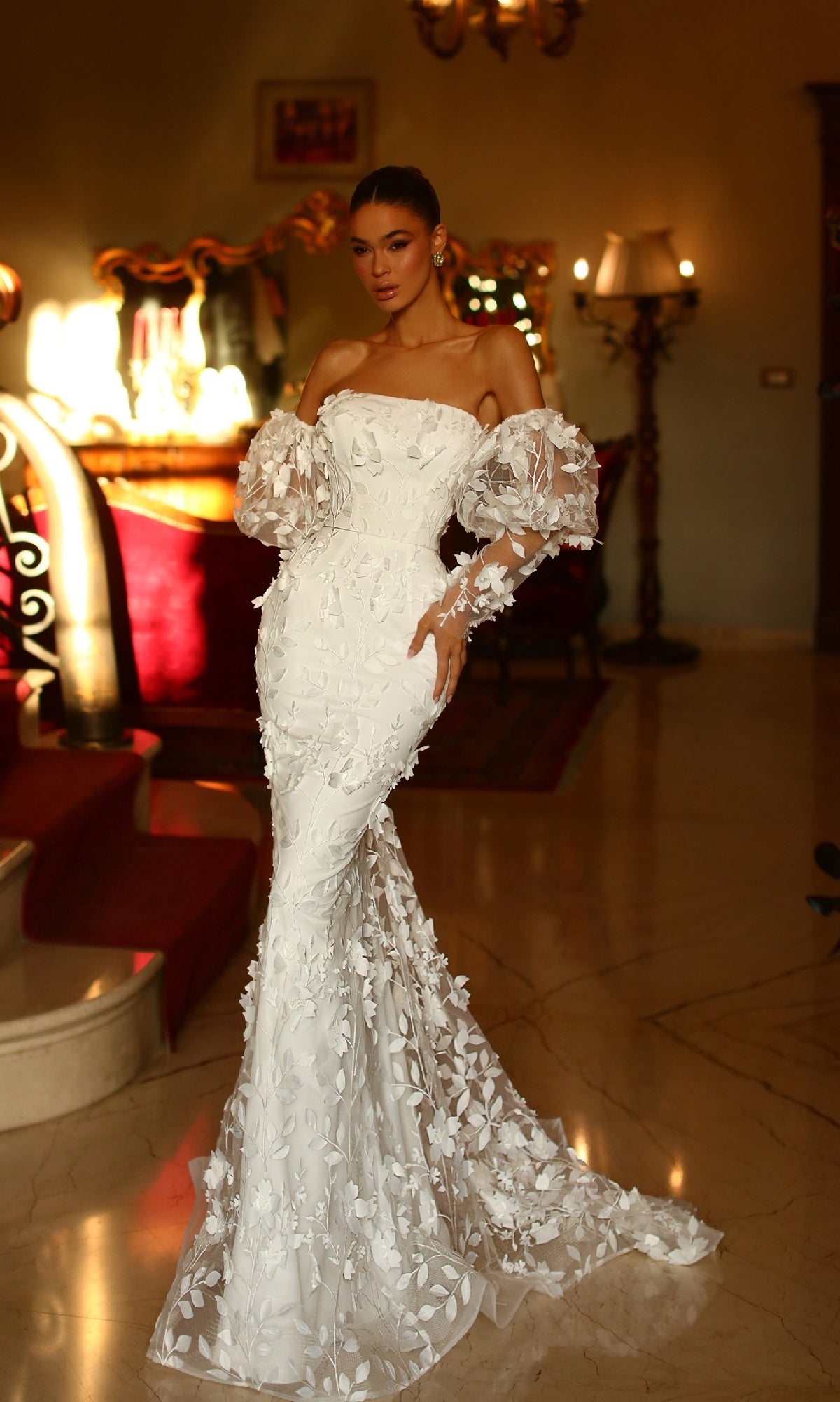 A woman stands indoors on a marble floor, wearing the Puff-Sleeve White Formal Gown: Tina Holly TW121W, featuring elegant white lace and floral appliqué, with warm lighting and ornate decor in the background.